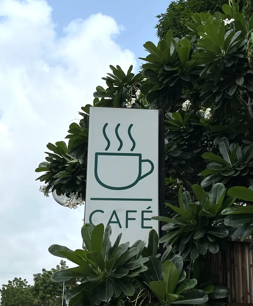 A painted café sign showing a steaming cup, mounted among frangipani leaves